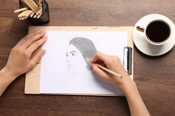 Ręce rysują ołówkiem portret kobiety na kartce; obok filiżanka kawy i pojemnik z ołówkami