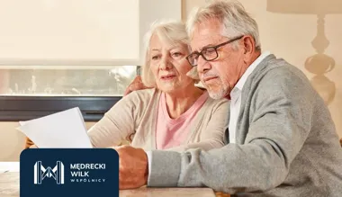 Starsza para siedzi przy stole i przegląda dokumenty, obok logo Mędrecki Wilk Wspólnicy