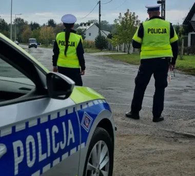 Dwóch policjantów w kamizelkach odblaskowych stoi przy policyjnym radiowozie na wiejskiej drodze.