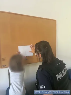 Policjantka w mundurze pomaga dziecku przyczepić rysunek do tablicy korkowej w pomieszczeniu.