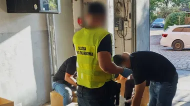 Funkcjonariusz Służby Celno-Skarbowej w żółtej kamizelce nadzoruje pracę mężczyzn sortujących towary w pomieszczeniu magazynowym przy otwartej bramie garażowej.