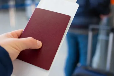 Dłoń trzyma bordowy paszport z włożonym do środka biletem lotniczym na lotnisku.