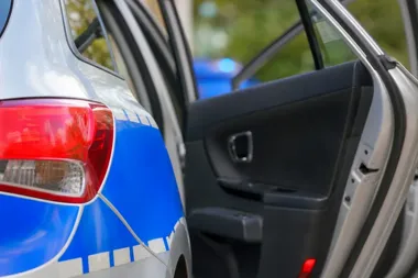 Fragment policyjnego radiowozu z otwartymi drzwiami, w tle błękitne światło.