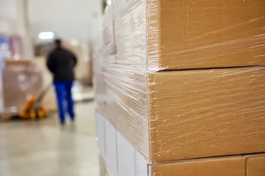 W magazynie stoją owinięte folią kartony na palecie, a w tle rozmazana postać pracownika w ciemnej kurtce i niebieskich spodniach idzie obok wózka widłowego.