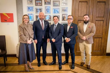 Grupa pięciu osób w strojach biznesowych pozuje do wspólnego zdjęcia w pomieszczeniu z obrazami i fotografiami na ścianie.